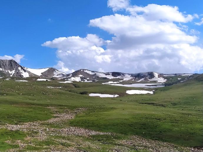 green grass field near snow covered mountains under blue and white sunny cloudy sky during daytime